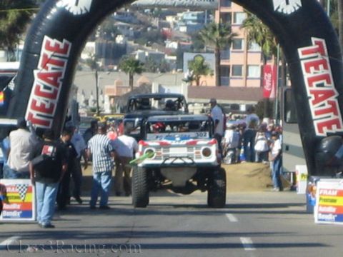 1990 Ford Bronco - Off-road race truck 302 approaches a Tecate-branded inflatable start arch with spectators lining a street start in a coastal town.