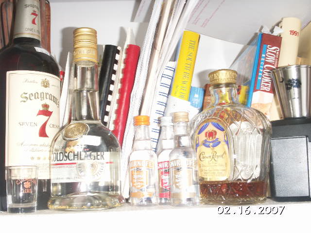 Several bottles of liquor, including Seagram's 7, Goldschlager, and Crown Royal, sit on a shelf with books and binders