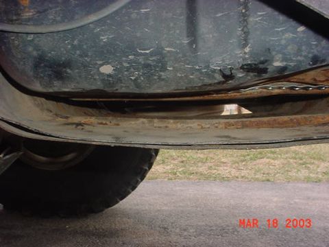 1980 Ford Bronco - Low-angle shot under a truck body showing a gas tank or skid pan with a surface-rusted mounting flange and a visible fastener, sitting just…