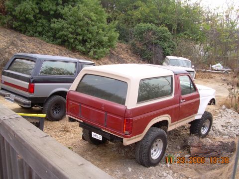 1990 Ford Bronco - Two older Ford Broncos parked on a dirt hillside, a gray one and a red-and-white lifted one viewed from the rear.