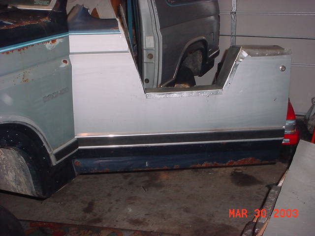 Cut white and blue Bronco door and rocker panels leaning against a stripped body in a garage