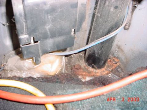 1980 Ford Bronco - Interior close-up of a black roll bar tube where it meets a rusty mounting foot on a corroded sheetmetal floor, with a fuel-tank or fender…
