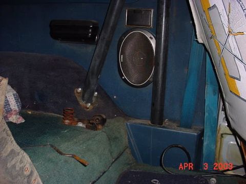 1980 Ford Bronco - Rear interior corner of a blue truck cab showing black-painted roll bar tubing bolted through the wheel-well tub, a large aftermarket coaxi…