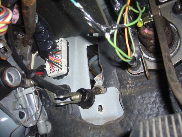 Under-dash view with white wiring connector, booted linkage rod, and black pedal edge.