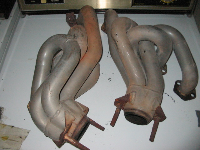 Two exhaust manifolds, one with rust-colored discoloration, resting on a white surface