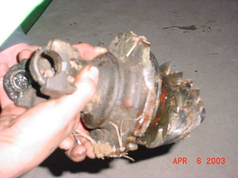 1980 Ford Bronco - Two hands holding a filthy, grease- and grass-caked axle U-joint/knuckle assembly with a torn rubber boot over a concrete floor. Rust and d…