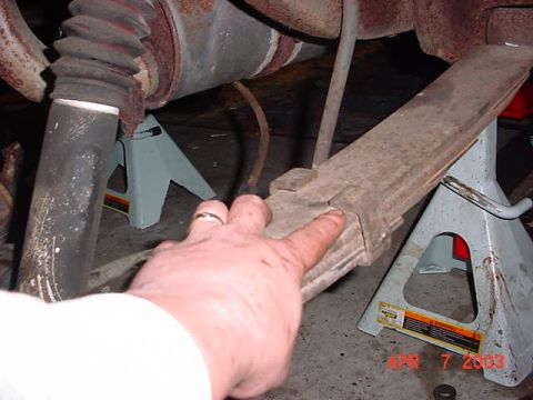 1980 Ford Bronco - Underbody shot with a hand pointing at a rusty leaf spring resting on a jack stand, taken beneath a vehicle supported on stands over a conc…