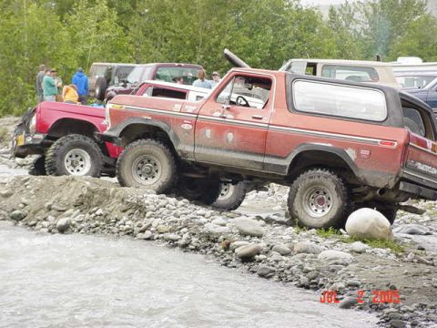 1978 Ford Bronco - Multiple off-road vehicles navigate a riverbed with rocky banks and trees in the background.