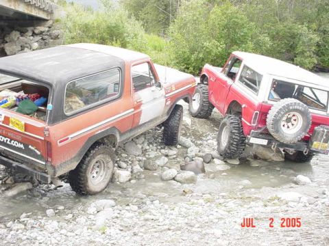 1978 Ford Bronco - Two Ford Broncos navigate a rocky stream bed under a bridge.