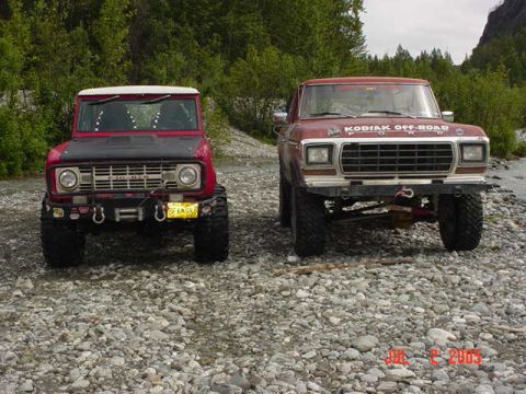 1978 Ford Bronco - Two SUVs are parked side-by-side on a rocky riverbed with trees in the background.