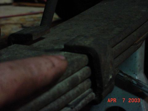1980 Ford Bronco - Dim close-up of a rusty multi-leaf spring pack held in a bench vise, with a thumb pointing at the retaining clip wrapped around the stacked…