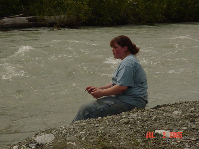 Person sitting on a gravel bank watching a fast gray glacial river