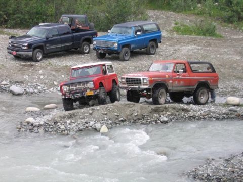 1978 Ford Bronco - Four-wheel-drive vehicles traverse a shallow river, with two red Ford Broncos and a blue SUV prominently featured.