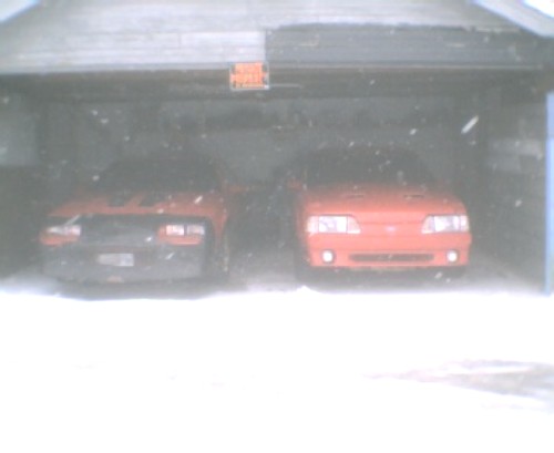 Two red cars parked inside a garage viewed from front, with snow falling outside