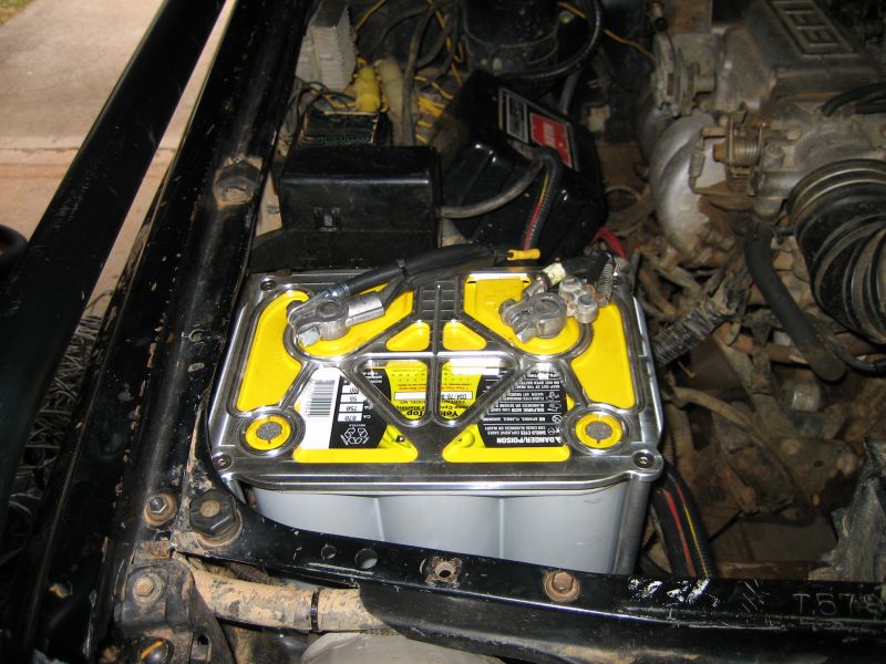 Yellow top car battery with metal hold-down bracket and connected terminals in engine bay