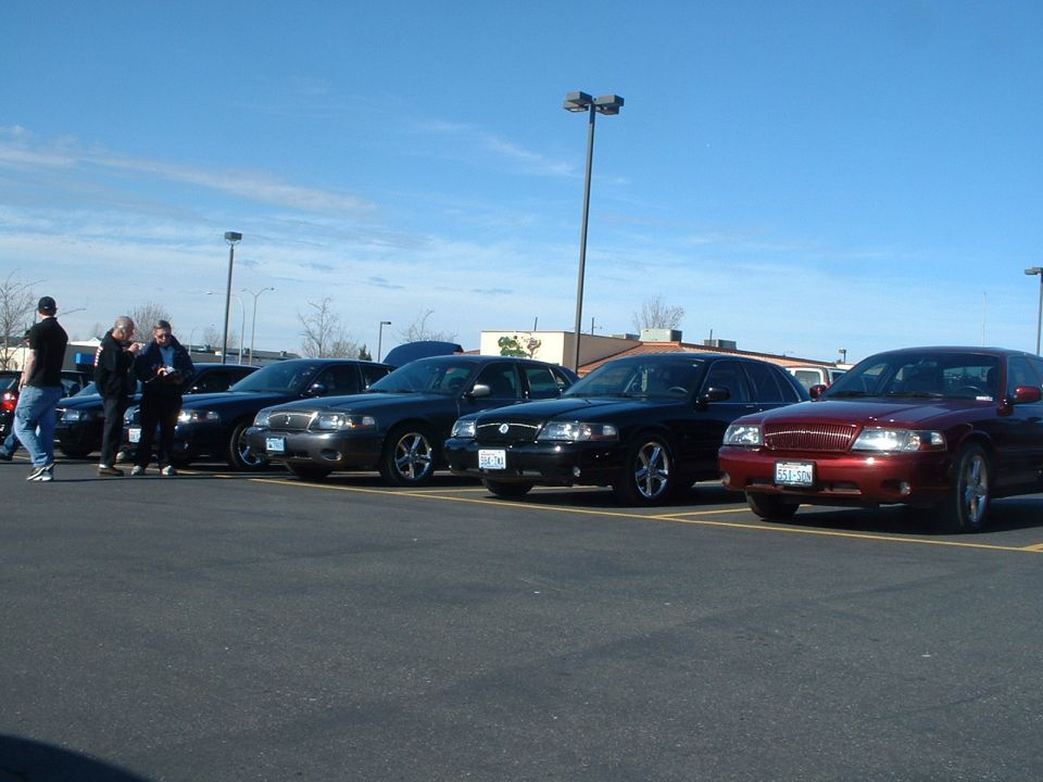 Several Ford Crown Victorias parked in a lot, one in red, others in black. Some people stand near the cars