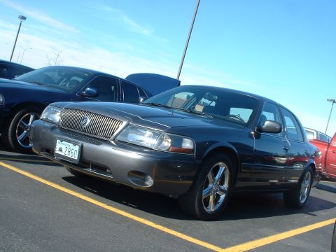 2000 Ford Crown Victoria - Two sedans are parked in a lot under a bright blue sky.