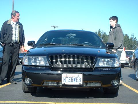 2000 Ford Crown Victoria - Two men stand near a black 2000 Ford Crown Victoria parked in a lot on a sunny day.