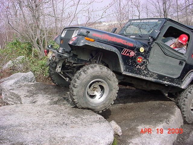 Muddy black Jeep Wrangler YJ crawling off a rock ledge on a wooded trail