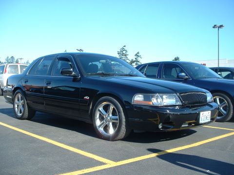 2000 Ford Crown Victoria - A black sedan with aftermarket wheels is parked in a lot under a clear blue sky.