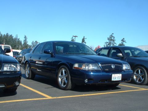 2000 Ford Crown Victoria - A blue Mercury Grand Marquis is parked in a lot, showing its distinctive grille and chrome wheels.