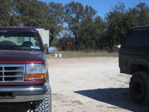 1996 Ford Bronco - Two Ford Broncos, one maroon and one black, are parked in a dirt lot with trees in the background.