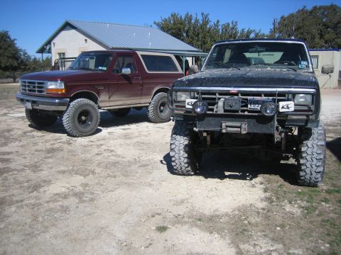 1996 Ford Bronco - Two Ford Broncos are parked on a dirt lot in front of a building on a sunny day.