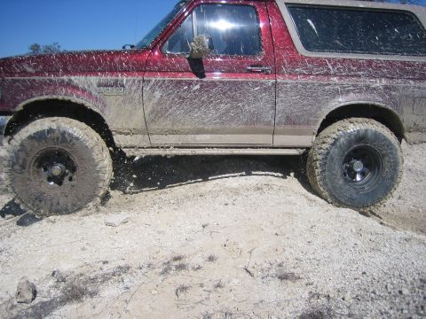 1996 Ford Bronco - A muddy red Ford Bronco navigates rough terrain with off-road tires visible.