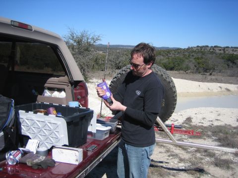 1996 Ford Bronco - A man examines a bottle while standing next to the open tailgate of a red SUV, which has a spare tire mounted on its rear.