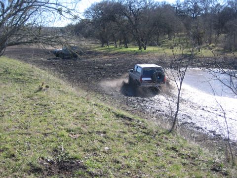 1996 Ford Bronco - Two SUVs navigate a muddy off-road course with trees and water visible.