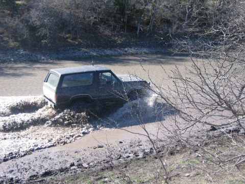 1996 Ford Bronco - A dark SUV navigates a muddy waterway, kicking up water and dirt.