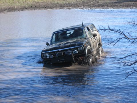 1996 Ford Bronco - Black SUV navigates through a water crossing, kicking up spray.