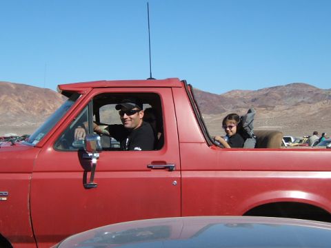 1988 Ford Bronco - A man and a child are in a red 1988 Ford Bronco, parked in a desert landscape with other vehicles visible.