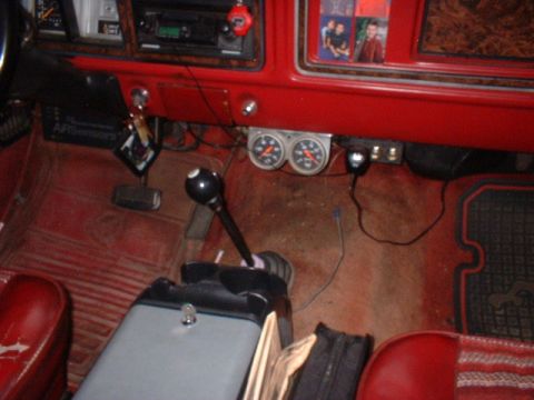1979 Ford Bronco - The interior of a vehicle shows a floor shifter, dual gauges, and a radio, with red seats and floor mats visible.