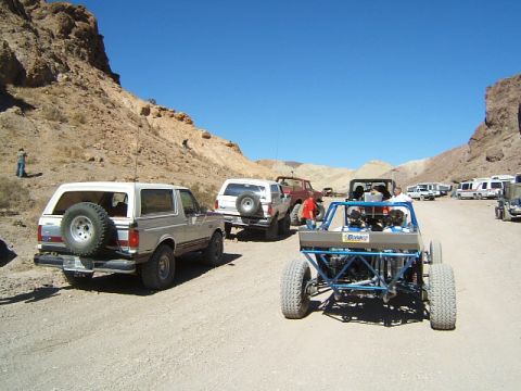 1988 Ford Bronco - A group of off-road vehicles are gathered in a desert area with rocky hills under a clear blue sky.