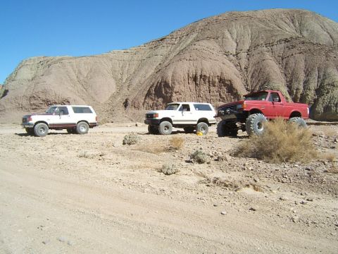 1988 Ford Bronco - Three off-road vehicles are parked on a dirt road in a desert landscape with a large, layered rock formation behind them.