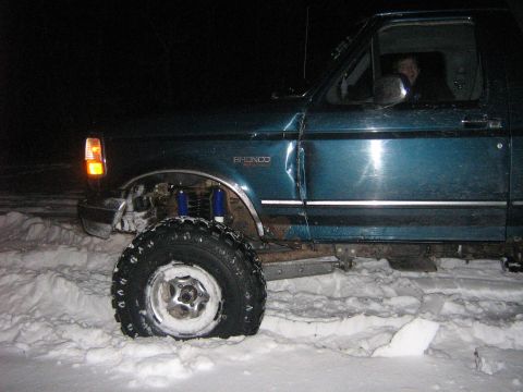 1995 Ford Bronco - A teal Ford Bronco with a lifted suspension is stuck in the snow, its front tire deflated.