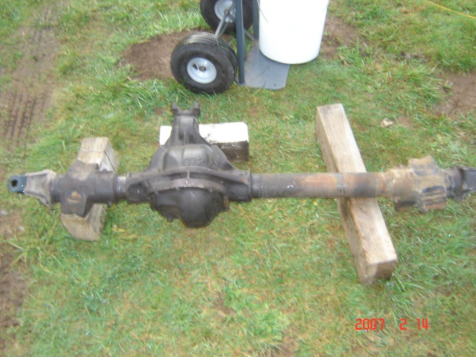 Vehicle rear axle assembly on wooden blocks outdoors. A dolly with wheels and a white bucket are in the background
