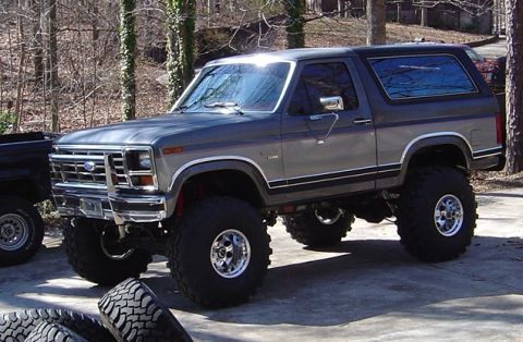 1986 Ford Bronco - A gray two-tone 1980s Ford Bronco full-size SUV lifted on large mud-terrain tires with polished aluminum wheels, chrome tube front bumper a…