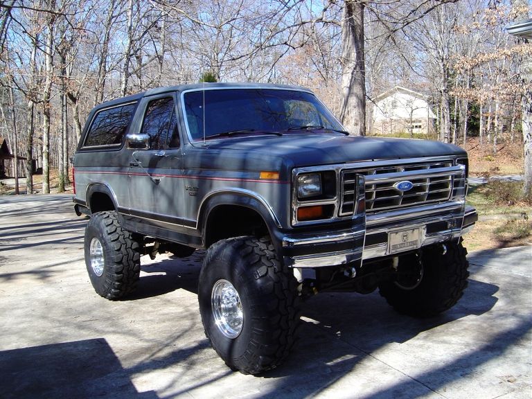 Lifted gray/black two-door Ford Bronco on big mud tires, front 3/4 view in a wooded driveway.