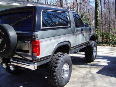 1986 Ford Bronco - Rear three-quarter view of a two-tone gray Ford Bronco full-size SUV lifted on large mud-terrain tires with polished aluminum wheels, showi…