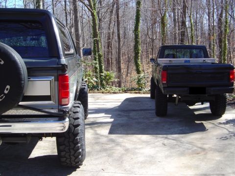 1986 Ford Bronco - Rear view of two lifted vehicles parked on a concrete driveway in a wooded area: a dark full-size SUV on the left with a rear-mounted spare…
