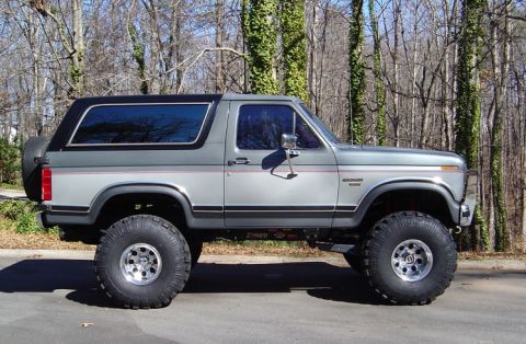 1986 Ford Bronco - Silver-gray two-door Ford Bronco SUV with black hardtop, shown in side profile, lifted on large off-road mud-terrain tires with polished al…