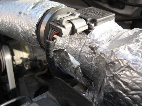 1994 Ford Bronco - Engine-bay close-up of a mass air flow sensor housing with its gray six-pin connector and loomed harness plugged in, sandwiched between int…