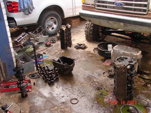 1987 Ford Bronco - Disassembled engine parts are scattered across a wet garage floor, with two trucks visible in the background.