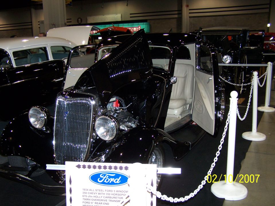 Black 1934 Ford 5-window coupe with white interior, hood open showing engine. Other cars in background