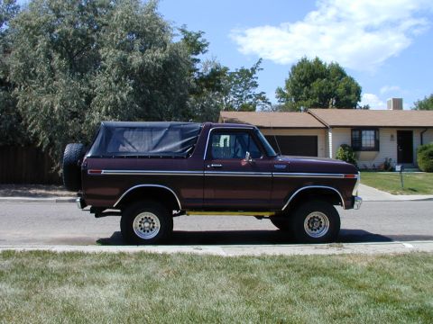 1979 Ford Bronco - Driver-side profile of a maroon full-size Ford Bronco with a black soft top, chrome bodyside molding and a rear-mounted spare tire, parked…