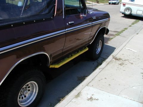 1979 Ford Bronco - A dark red Ford Bronco with yellow step bars is parked on the side of a street next to a camper trailer.