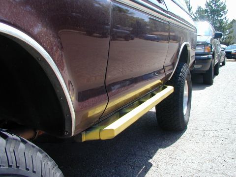1979 Ford Bronco - Low close-up along the passenger side of a maroon Ford SUV showing a yellow tube rock slider / side step bar mounted under the rocker betwe…