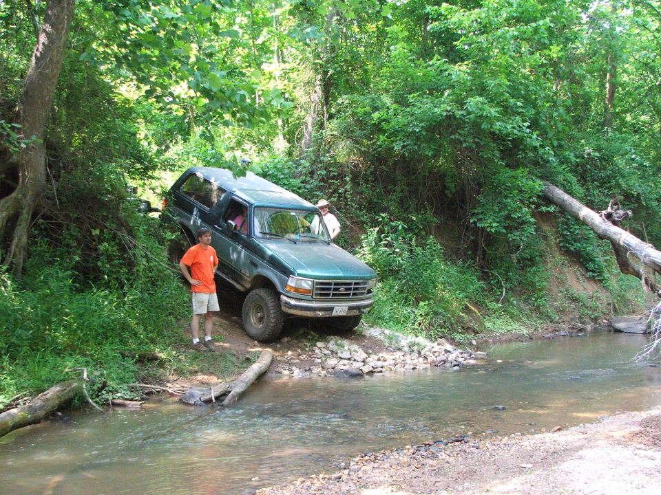 Green Ford Bronco on a dirt path next to a stream with two people nearby
